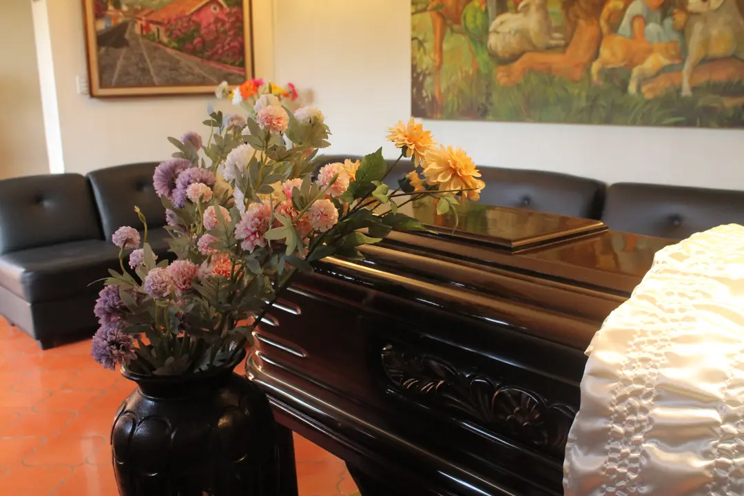 Ataúd de madera oscura con un arreglo de flores, en una sala con sillones y cuadros