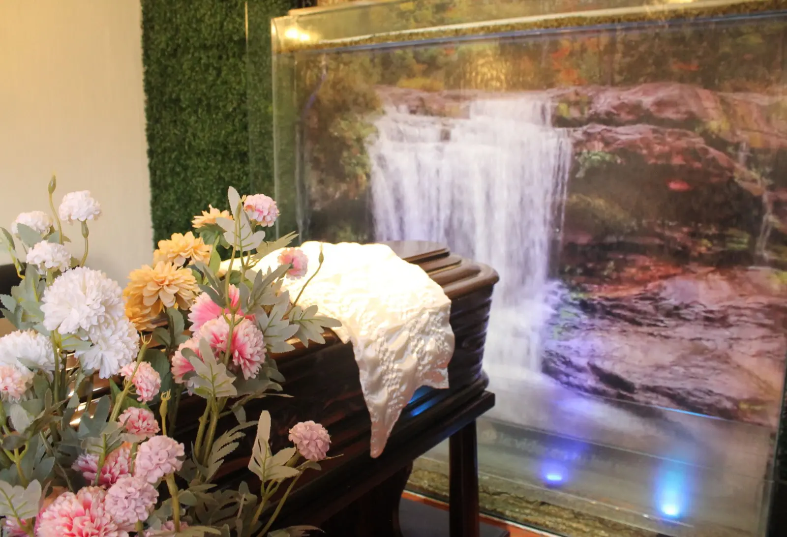 Capilla de Hosanna: ataúd con paño blanco y un arreglo de flores frente a una cascada de agua iluminada