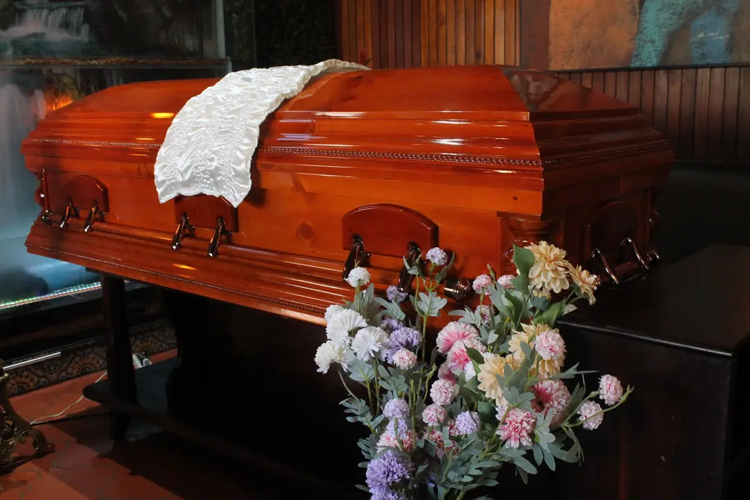Ataúd de madera barnizada con herrajes y un paño blanco, junto a un arreglo de flores